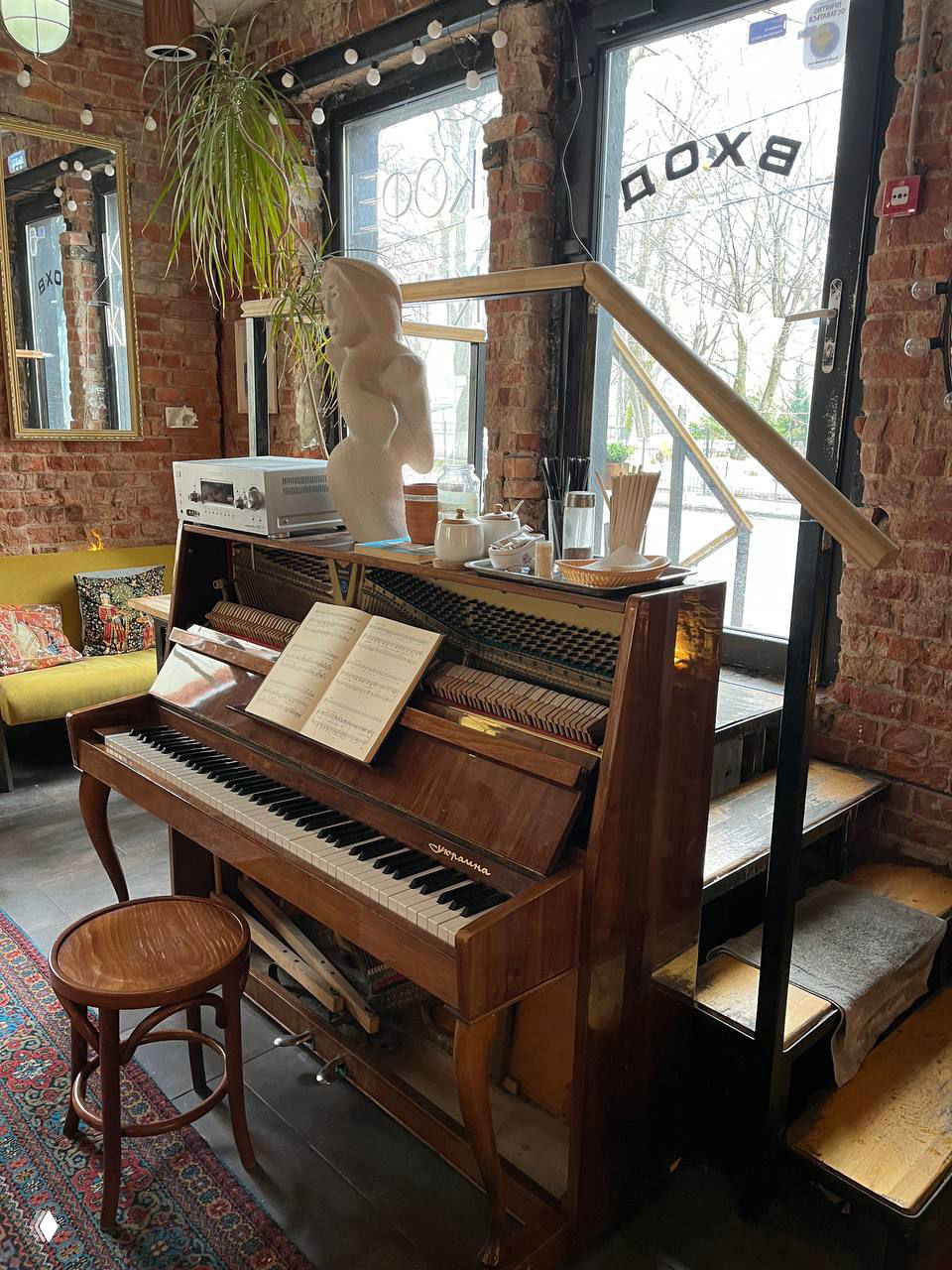 Декоративное фортепиано в пространстве кафе арт‑хостела «Толстой», раскрытые ноты на подставке и лестница у входа на фоне витрины.