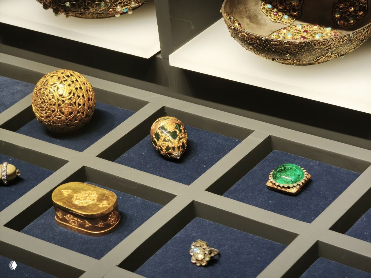 Несколько экспонатов Фаберже — яйца, шкатулки и броши на тёмно‑синей подложке; приглушённый музейный свет подчёркивает текстуры и деталь.