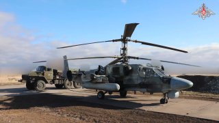 Боевой вертолёт на стоянке у укрытия в открытой полевой местности; виден весь фюзеляж и эмблема в правом верхнем углу кадра, нейтральный ракурс.