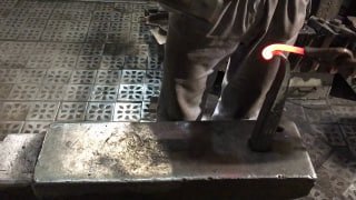 Кадр кузнеца у наковальни: раскалённый прут в тисках, видна рабочая зона и наковальня — процесс ручной ковки в мастерской.