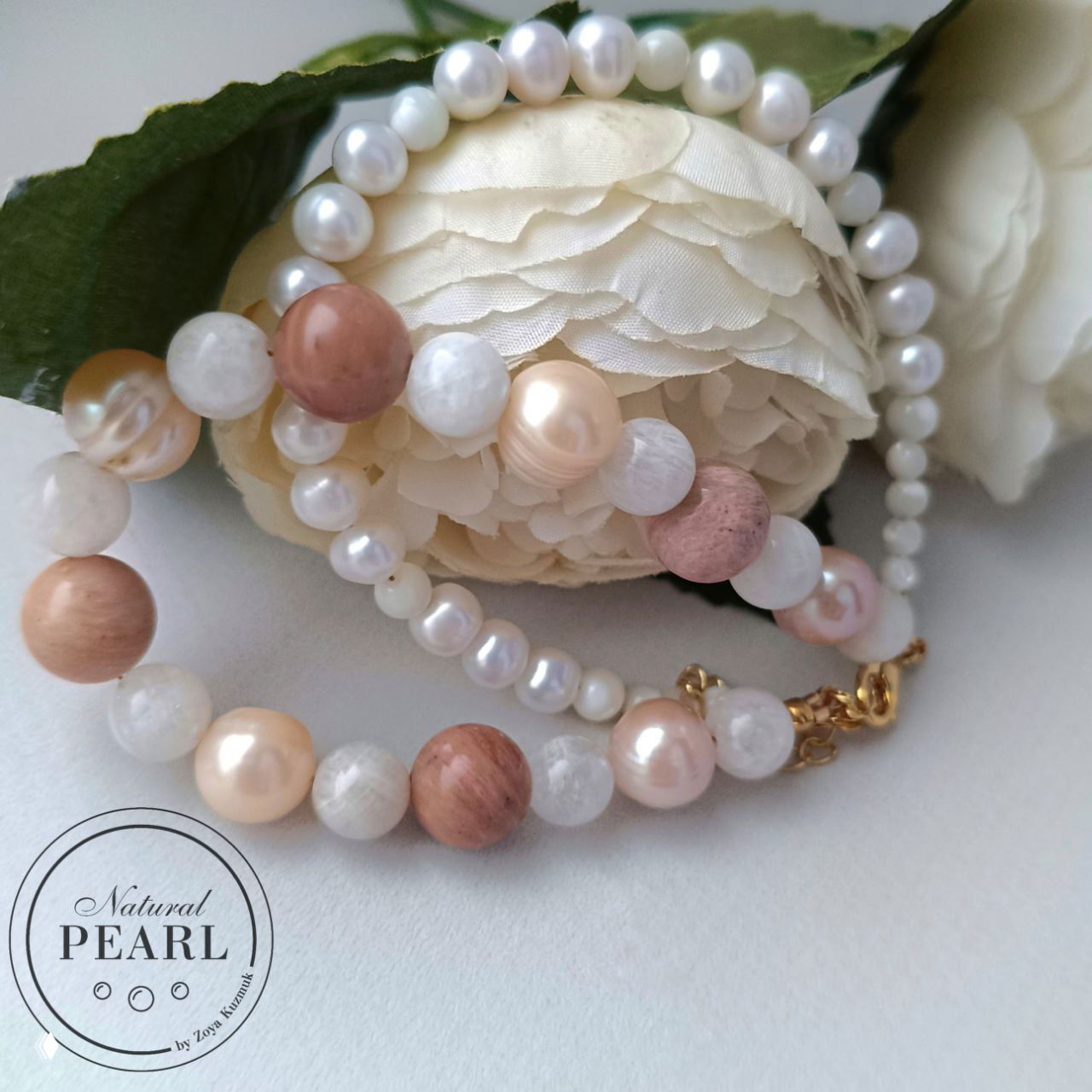 Браслет уложен около искусственного цветка: жемчуг, перламутр и розовый родонит сочетаются с золотистой застёжкой на светлом фоне.