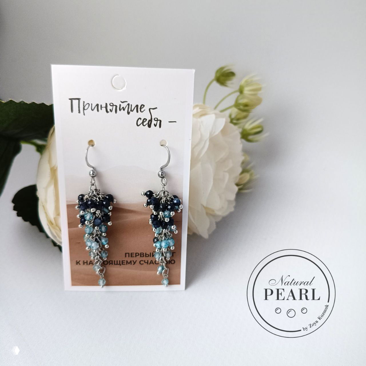 Вид спереди: серьги‑грозди на фирменной карточке, рядом белые декоративные цветы — фотография для портфолио авторских украшений.