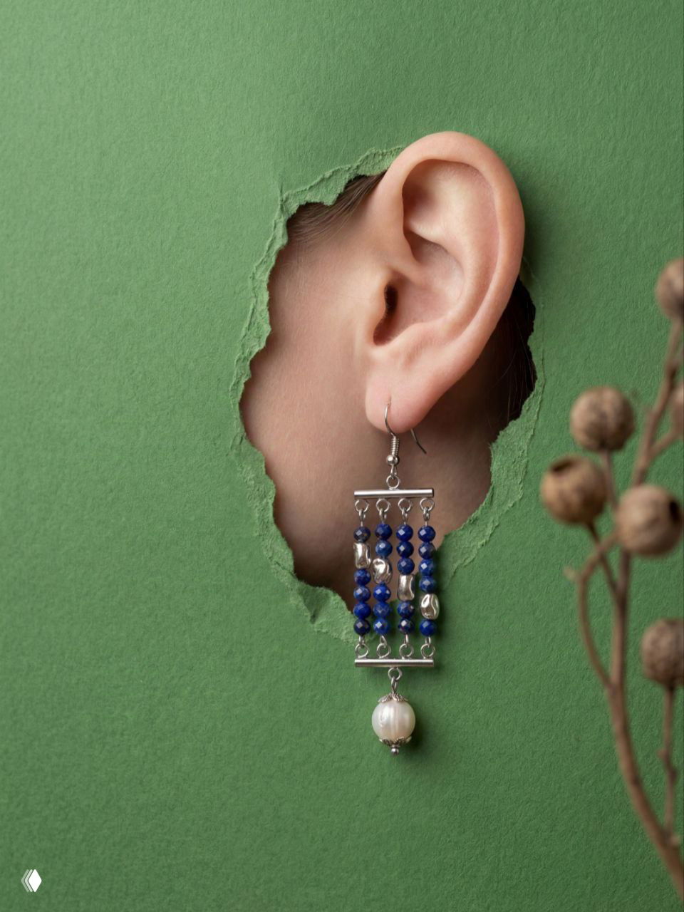 Серьга в ухе, показанная через прорезь зелёной бумаги: сочетание тёмно-синих лазуритовых бусин, серебристых деталей и жемчужного подвеса.