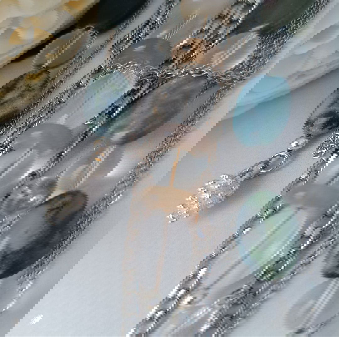 Крупный план украшений: прозрачные и зелёные бусины, цепочки и маленькая шишка-подвеска на светлом фоне — детали изделия.