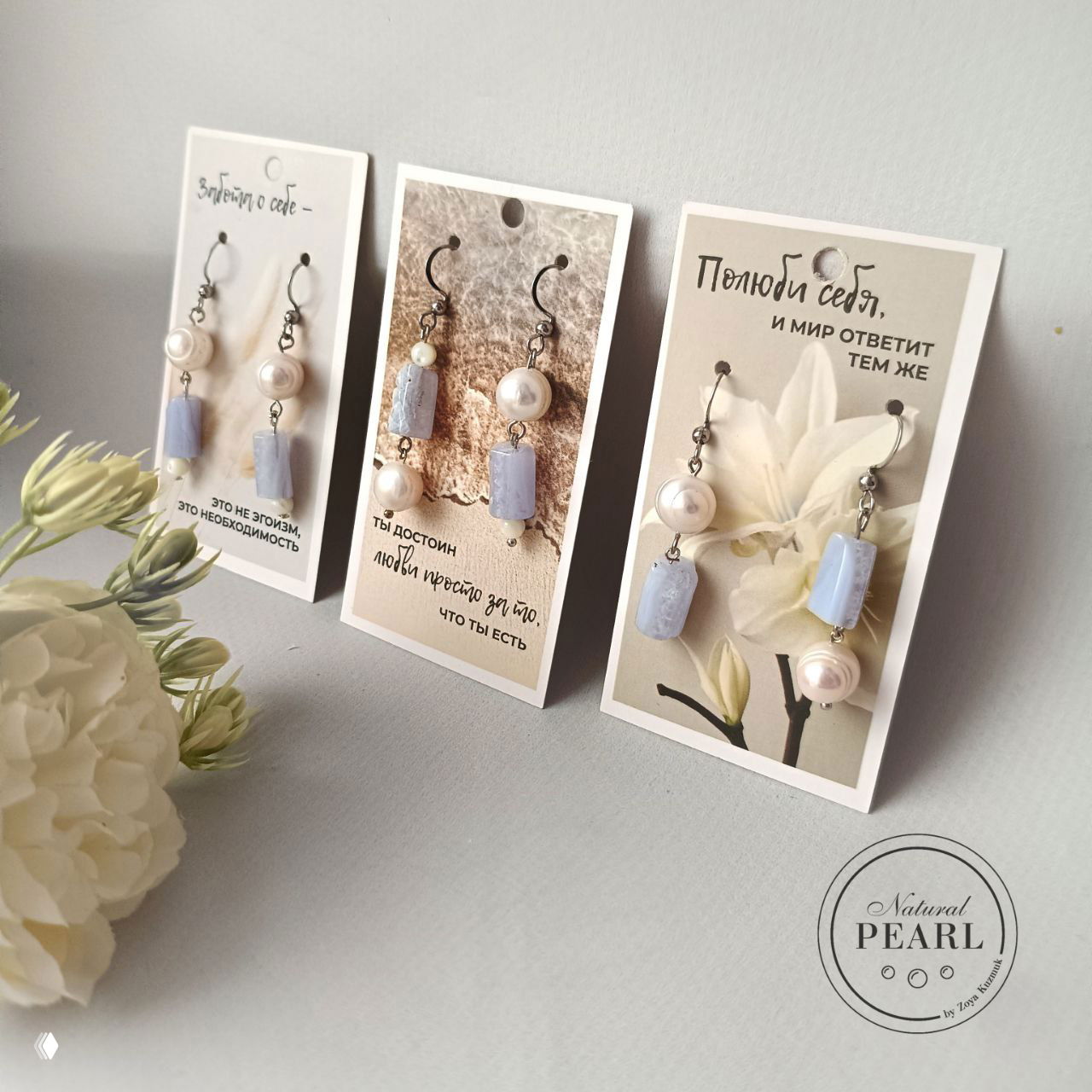 Три карточки с парой серёг: голубой сапфирин в цилиндрической огранке и белые жемчужины на фоне декоративных карточек и цветка.