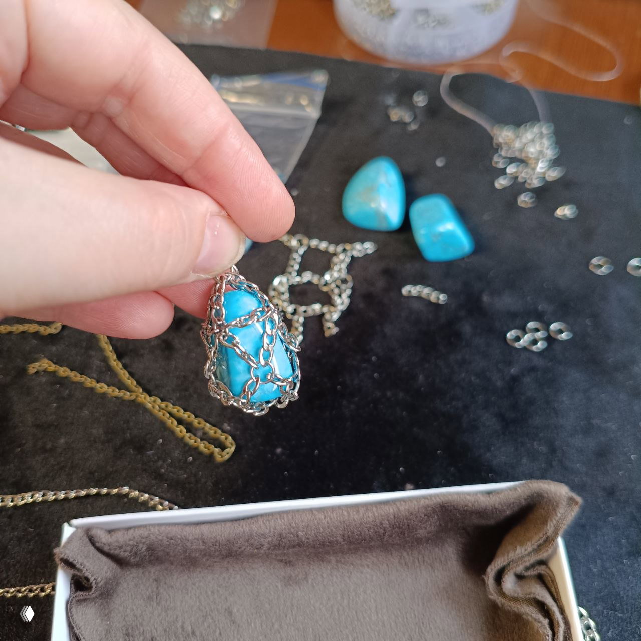 Рука мастера держит ярко-голубой камень в цепочной оплётке на коврике для работы: подготовка к сборке и закреплению украшения.