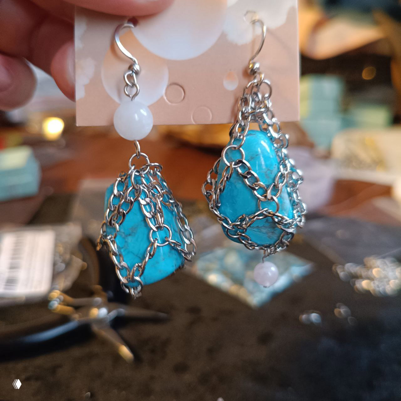 Крупный план серёг: ярко-голубые говлитовые камни в металлической цепочной оплётке на рабочем столе мастера, видны инструменты и фон.