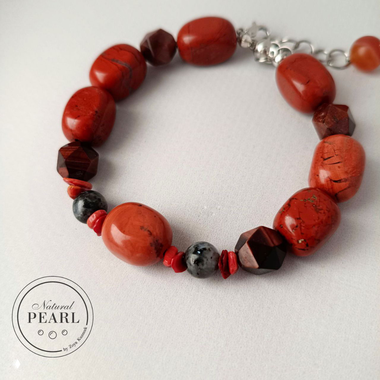 Общий вид браслета на светлом фоне: красные камни коралла/яшмы образуют окружность, видны гранёные тёмные элементы и серые ларвикитовые бусины, фирменный логотип в кадре.