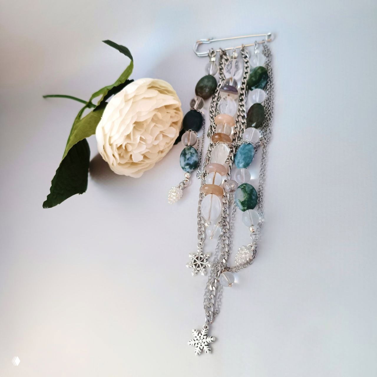 Композиция браслетов и подвесок: плоские и круглые бусины индийского агата и прозрачного кварца на серебристых цепочках рядом с белым цветком, студийная съёмка.