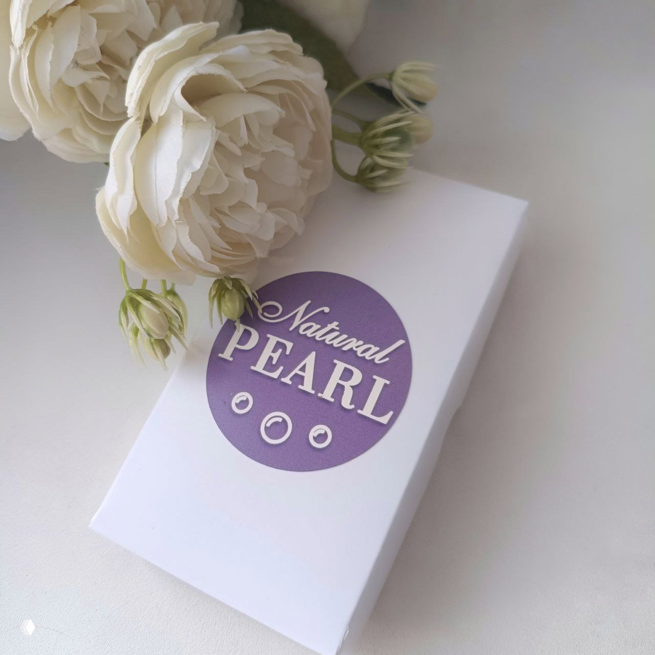 Фирменная белая коробка Natural PEARL с сиреневым логотипом на столе рядом с декоративными белыми цветами — упаковка украшения от автора.
