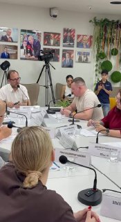 Участники круглого стола за белым столом с микрофонами и табличками: обсуждается ситуация на рынке банкротства физических лиц, видны камеры и техника.