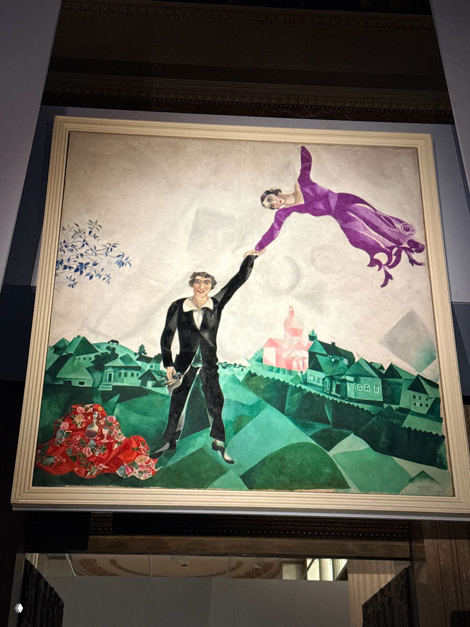 Картина Шагала: мужчина стоит на земле, женщина в пурпурном платье парит над ним, зелёный пейзаж и стилизованная архитектура на заднем плане.