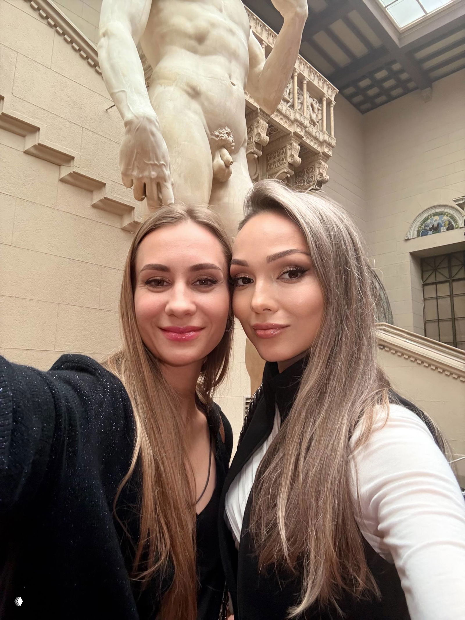 Две женщины делают селфи в музейном зале на фоне большой классической мраморной скульптуры с оголённой мужской фигурой и архитектурой интерьера.