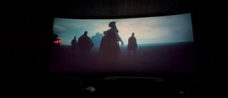 Фото экрана в тёмном кинотеатре: широкая панорама с силуэтами фигур на горизонте, передний план — тёмные кресла и приглушённый зал.