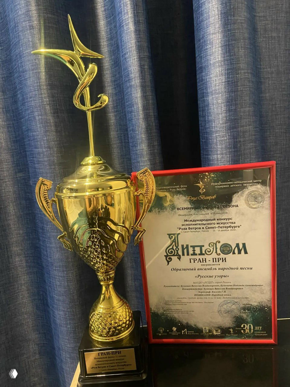 Крупный план золотого кубка и оформленного диплома с надписью «Гран‑при», выставленных на столе на фоне синих штор сцены