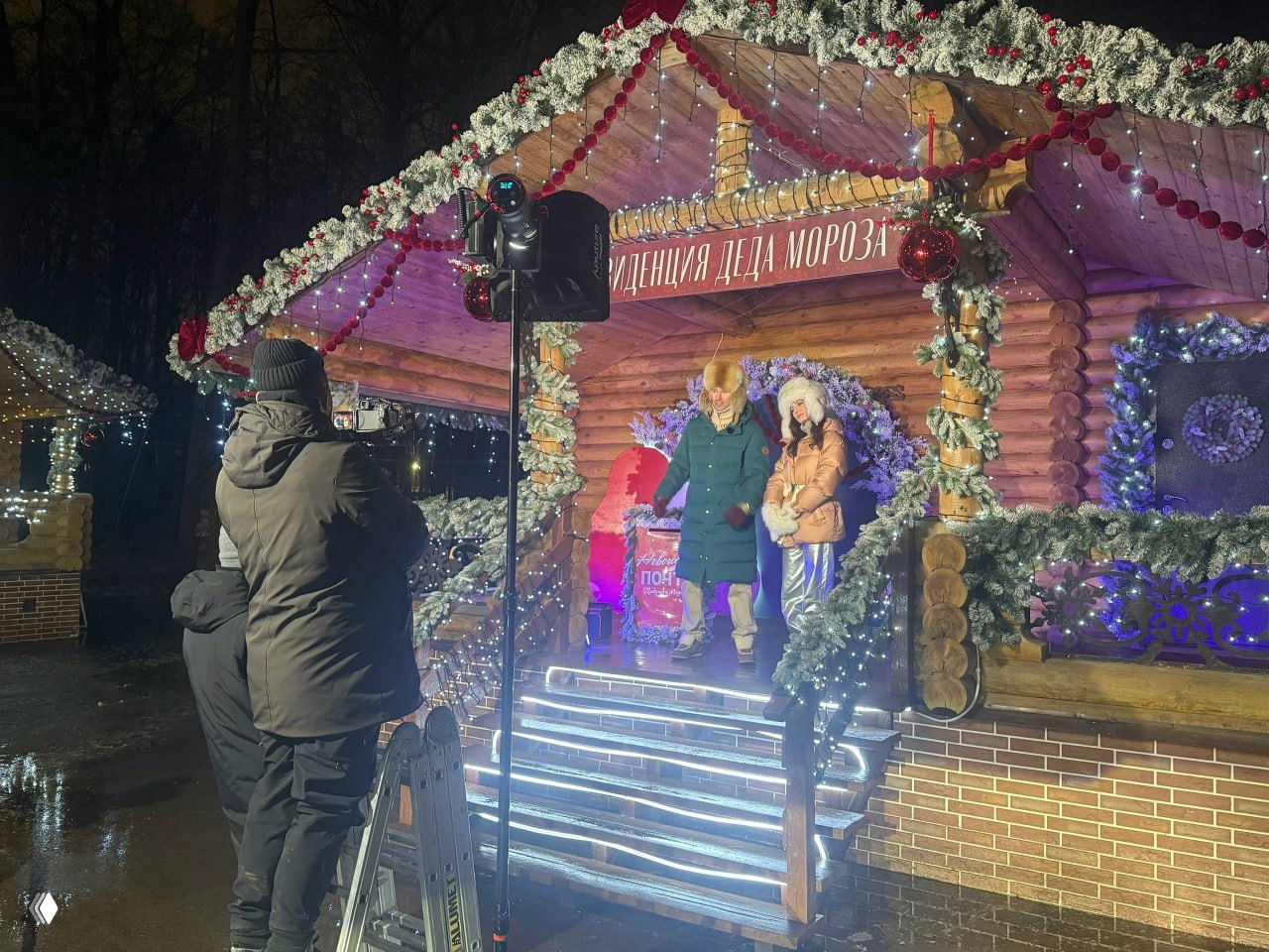 Ночная съёмка у украшенной резиденции Деда Мороза: операторы, свет и ведущие на ступенях светящегося павильона в Лесопарке.