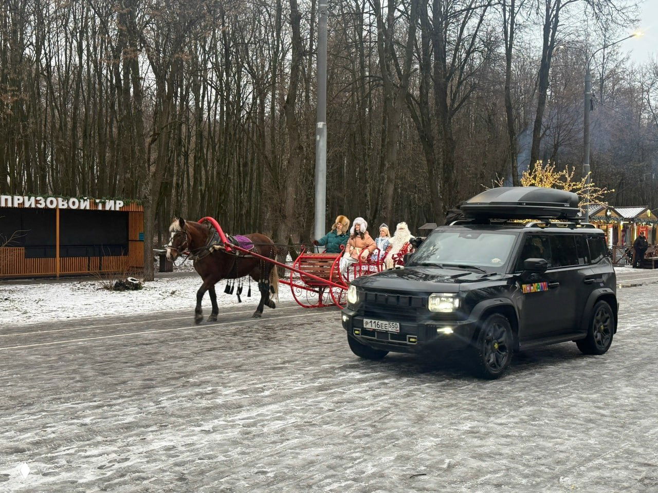 Конная повозка с Дедом Морозом и Снегурочкой и сопровождающий автомобиль проезжают по заснеженной дорожке Рязанского Лесопарка во время съёмок.