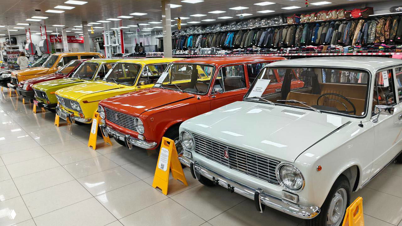 Ряд разноцветных советских легковых автомобилей ВАЗ в выставочном зале: белые, жёлтые, оранжевые и красные кузова на светлом полу, таблички рядом, Рязань.