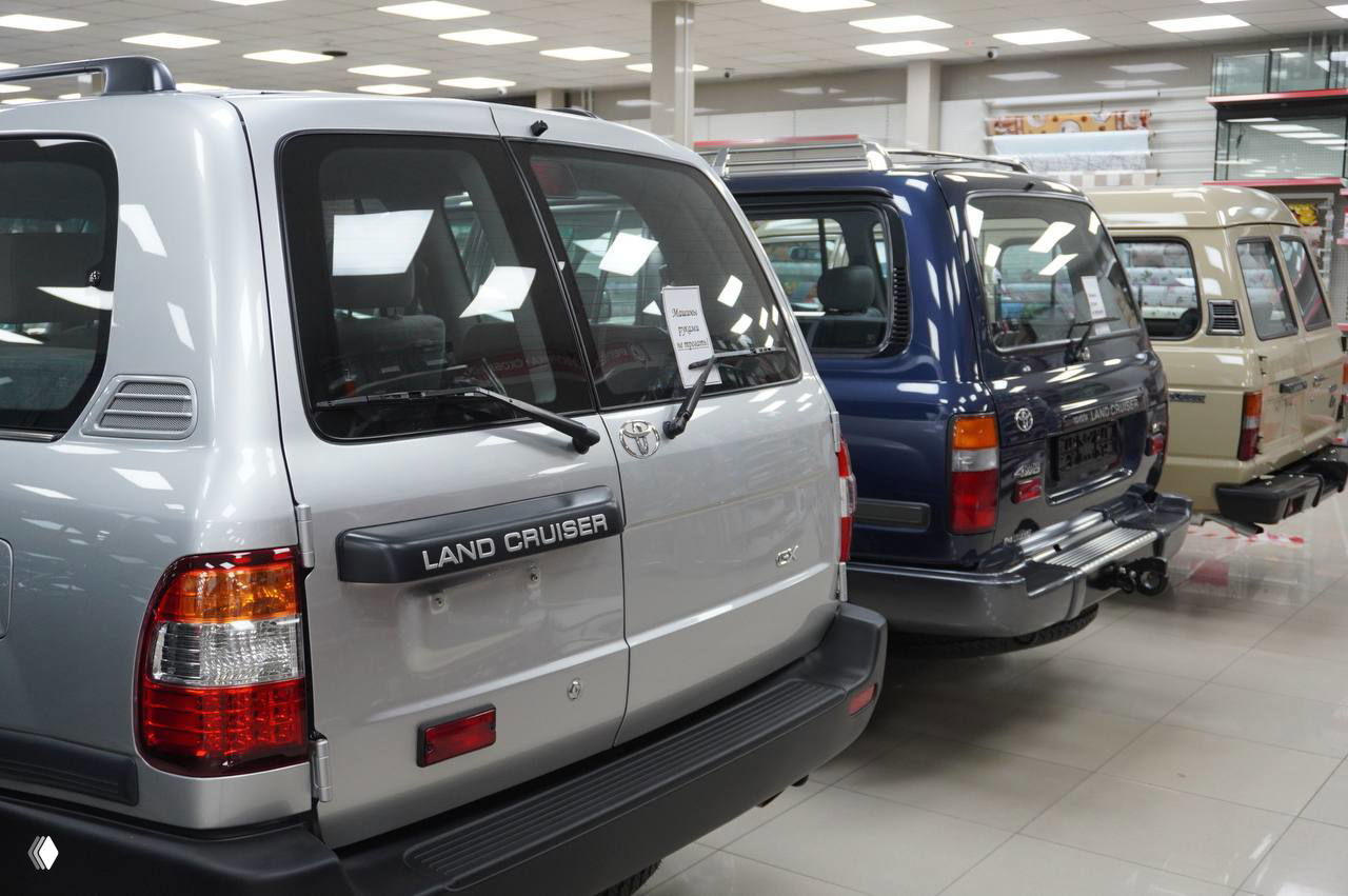 Задние части серого Land Cruiser и синего внедорожника в ряду ретроавтомобилей, вид на надписи и стоп‑сигналы, выставочный пол и информационные таблички на фоне, Рязань.