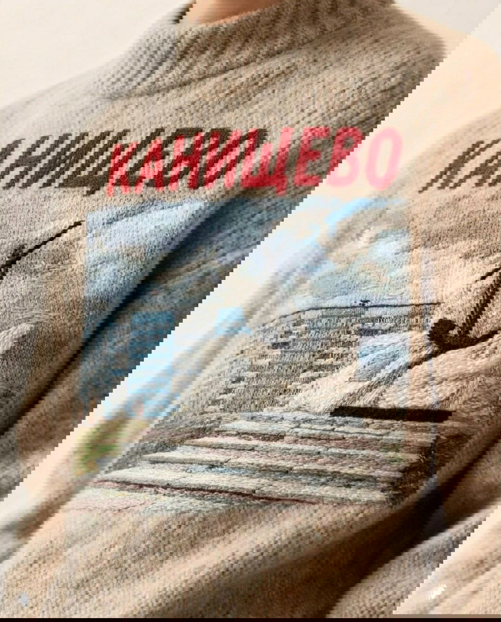 Светлый бежевый свитер с вышитой надписью «КАНИЩЕВО» и изображением монумента с якорём на фоне многоэтажной застройки — городской монументальный сюжет.