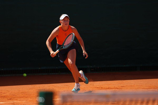 Динамичный кадр: теннисистка выполняет удар на грунтовом корте, видна ракетка, мяч и характерное оранжевое покрытие корта.