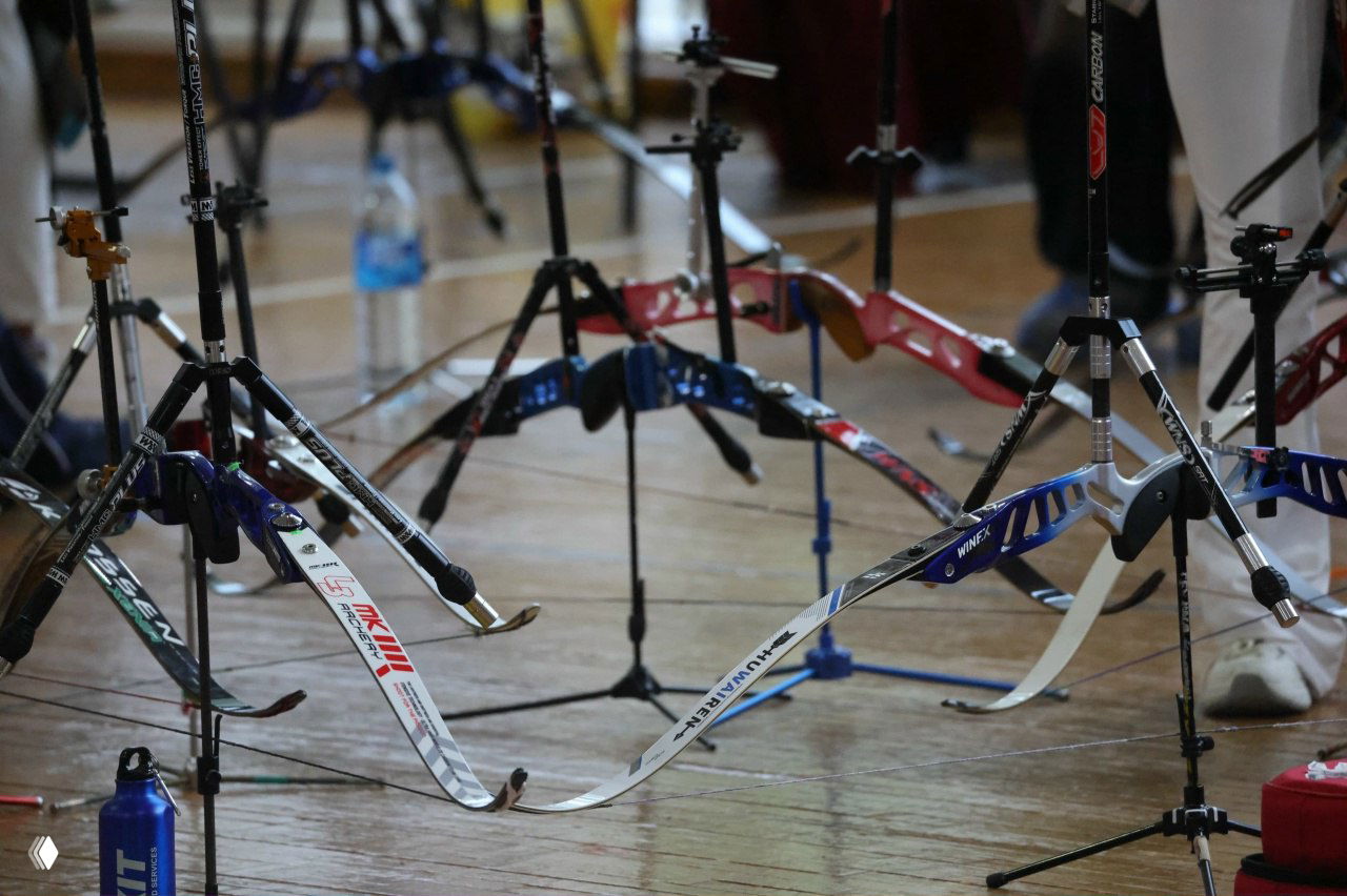 Ряды классических и рекурсивных луков на подставках в зале соревнований «Буревестник», видны стойки, кейсы и спортивная экипировка.