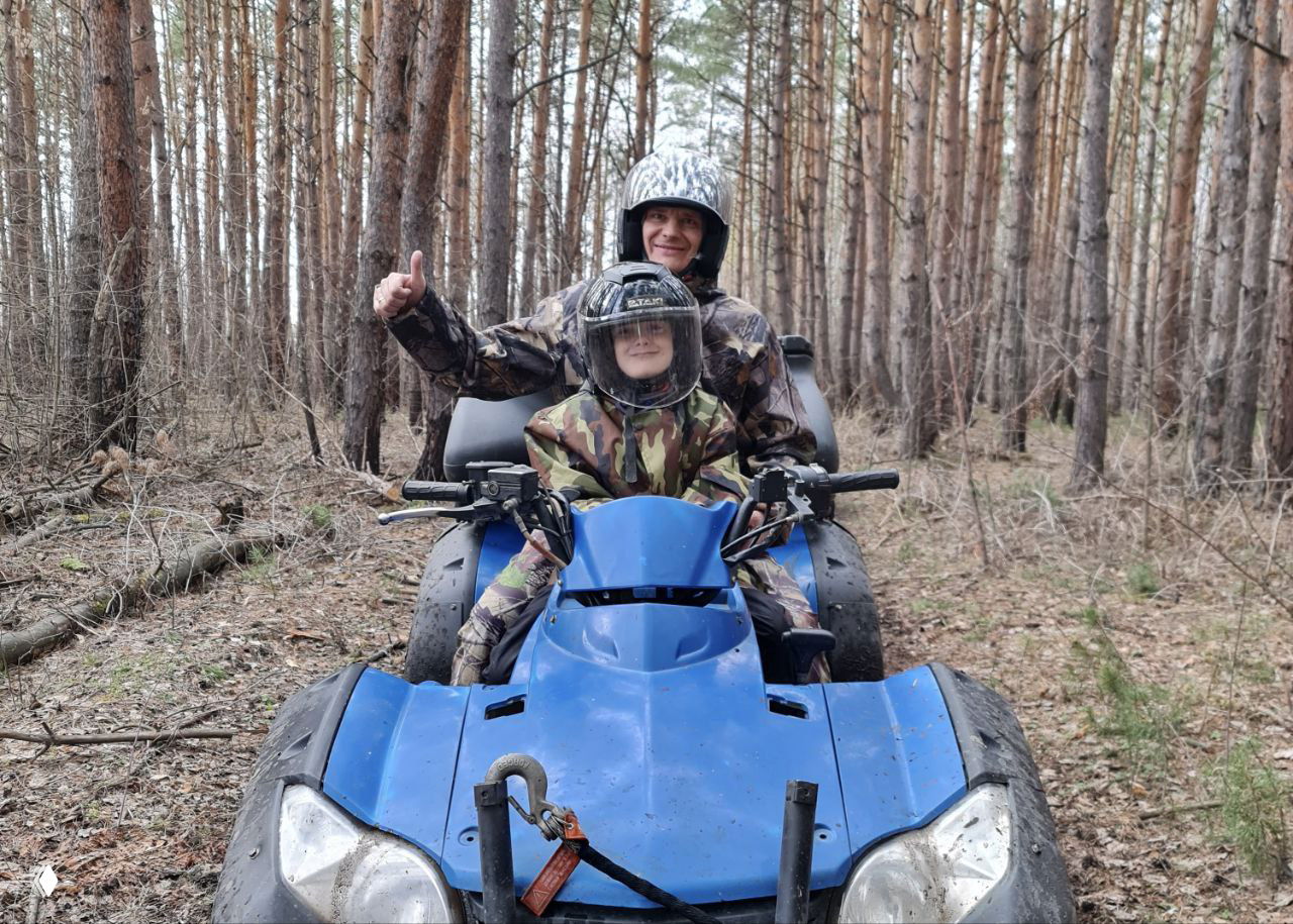 Отец с ребёнком на синем квадроцикле в сосновом лесу, оба в шлемах; сцена безопасности и семейного катания на маршруте «Тест-драйв» в прокате.