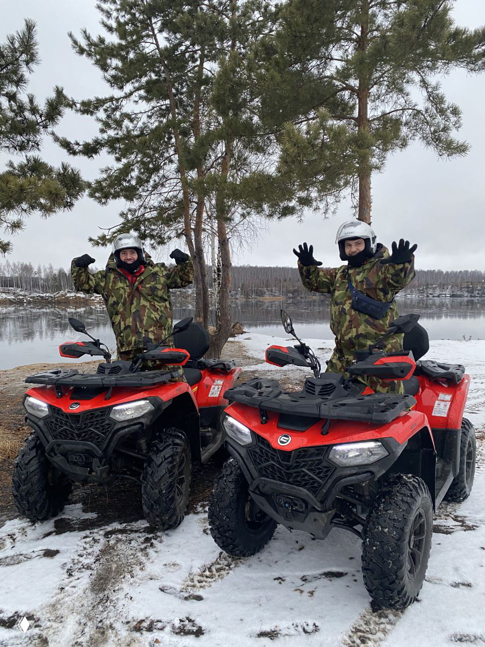 Два красных квадроцикла на заснеженном берегу озера, рядом стоящие водители в камуфляжной экипировке показывают жесты приветствия; зимний вариант маршрута проката техники.