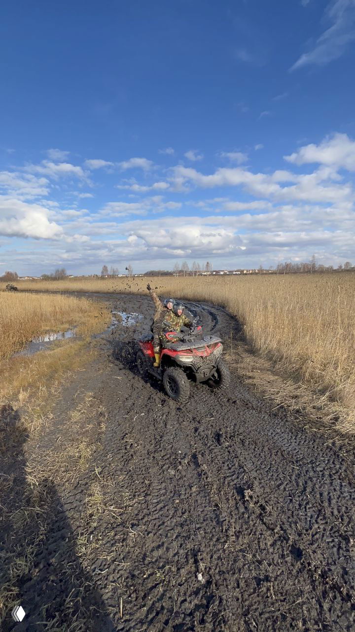 Красный квадроцикл едет по вязкой грязевой дороге под синим небом, водитель и пассажир поднимают руки — пример бездорожья и весёлого адреналинового участка маршрута.