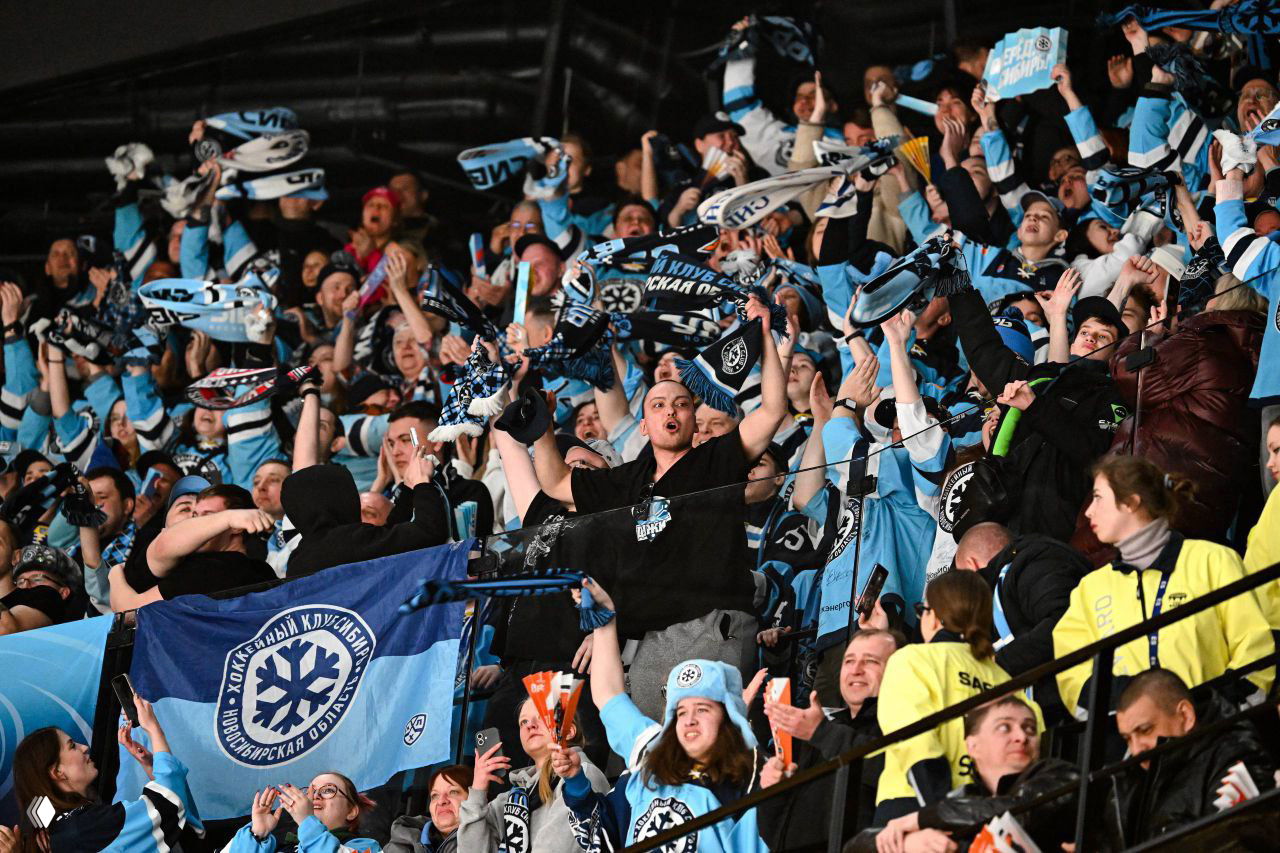 Плотные трибуны болельщиков ХК Сибирь в сине‑белой атрибутике: шарфы, флаги и скандирование даже при поражении.