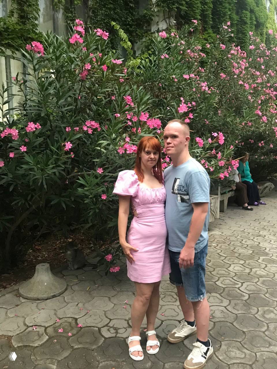 Пара стоит у цветущих кустов на аллее санаторной территории; прогулка по территории, видны зелень и дорожка.