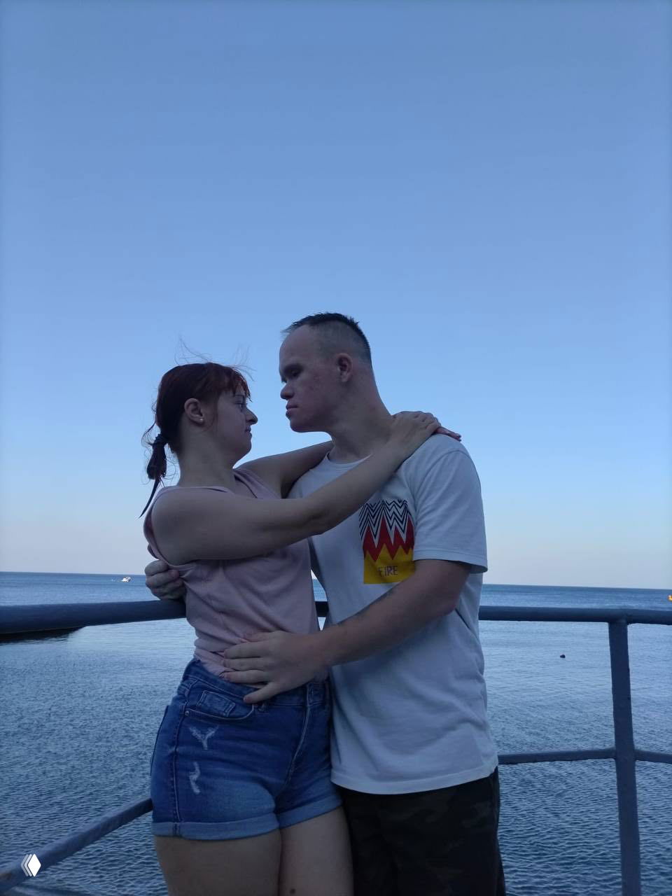 Пара обнимается на набережной у моря вечером, на заднем плане спокойный горизонт и перила; романтичный кадр на фоне моря.