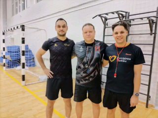Тренеры и организаторы турнира позируют в спортивном зале: три человека в спортивной форме на фоне ворот и гимнастического инвентаря, виден логотип «Синдром любви» на футболке.