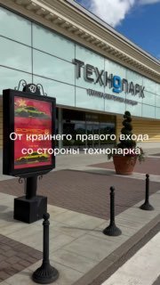 Кадр из видео от стороны Технопарка: вход с вывеской «ТЕХНОПАРК», рекламный пилон и элементы урбанистического оформления у фасада аутлета.