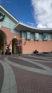 Кадр видеомаршрута: центральная зона Outlet Village Пулково у главного входа, арки, витрины и открытая пешеходная зона под голубым небом.
