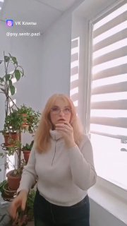 Женщина-психолог в светлом кабинете у окна с жалюзи, задумчиво держит руку у подбородка; на фоне комнатные растения, домашняя атмосфера.