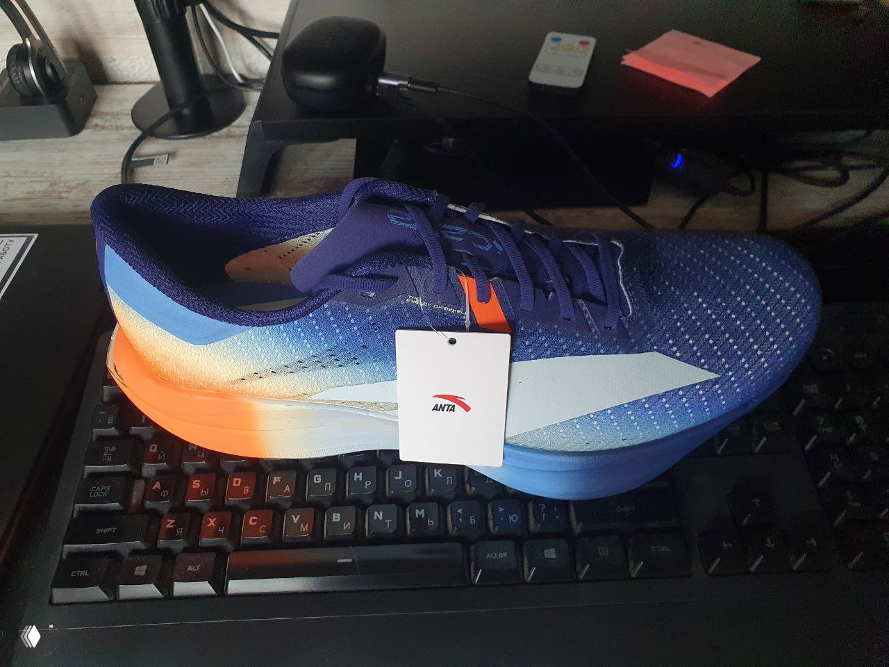 Синий и оранжево-белый беговой кроссовок ANTA C202 7 с прикреплённой биркой, вид сбоку, размещён на компьютерной клавиатуре в комнате как предпросмотр анонса.
