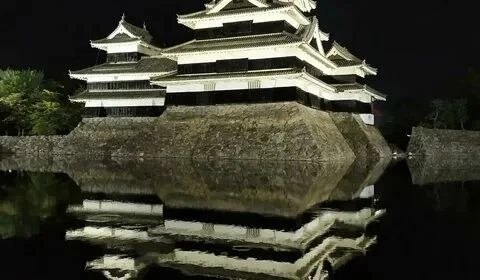 Ночной вид на стену и башни замка Мацумото с их отражением в спокойной воде рва, контраст света и темноты.