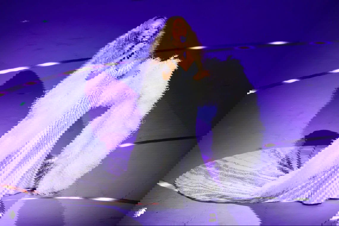 Солистка в длинном сверкающем белом платье с перьевыми элементами поёт на сцене под точечные световые эффекты и фиолетовый фон.