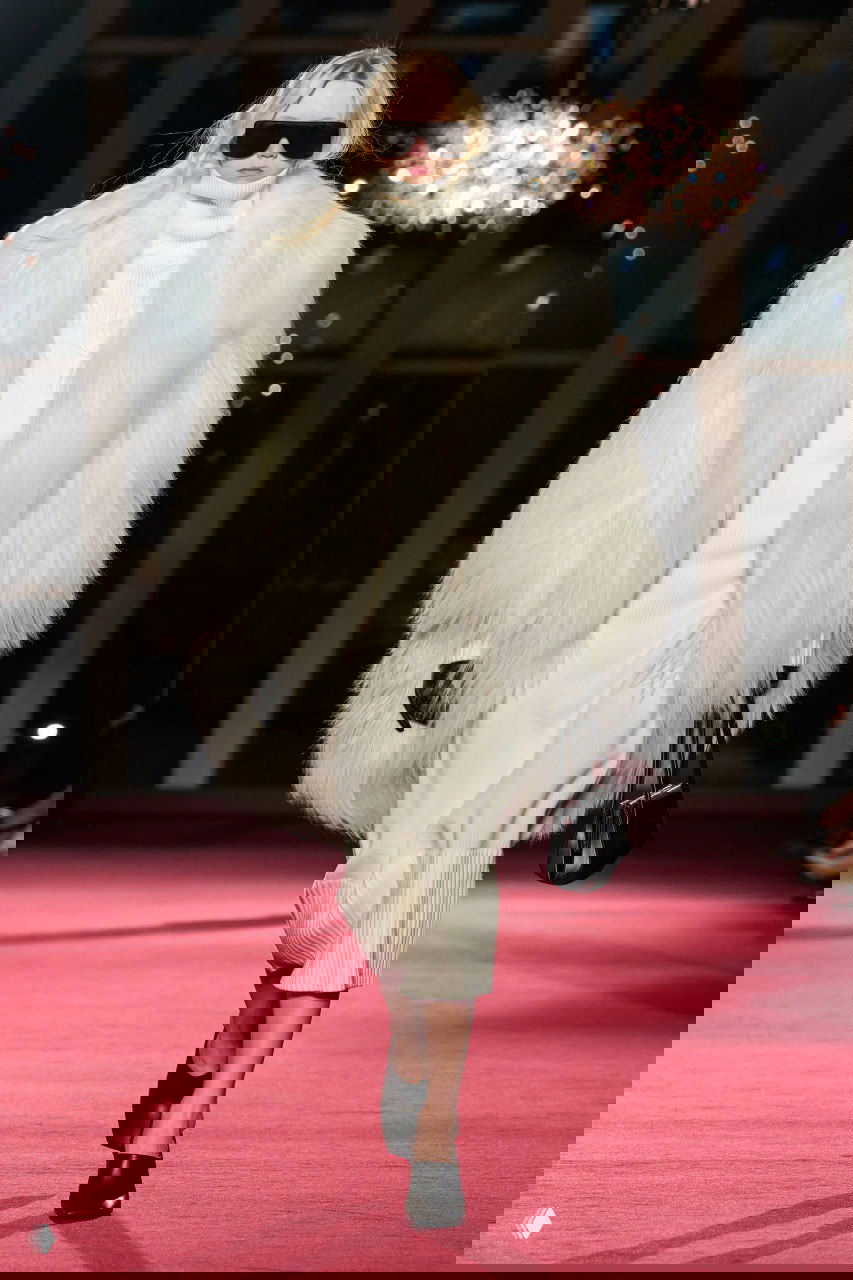 Модель в объёмной белой шубе и вязаном платье на подиуме Michael Kors в Метрополитен-опере; солнцезащитные очки и сумка как часть образа.