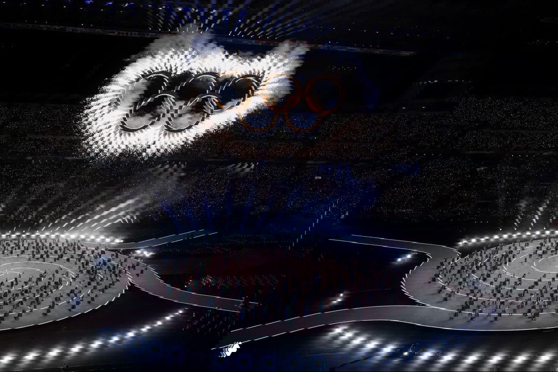 Широкий план арены: участники выстроены на спиральной круговой сцене, над ними горят кольца и идут пиротехнические эффекты в темноте стадиона.