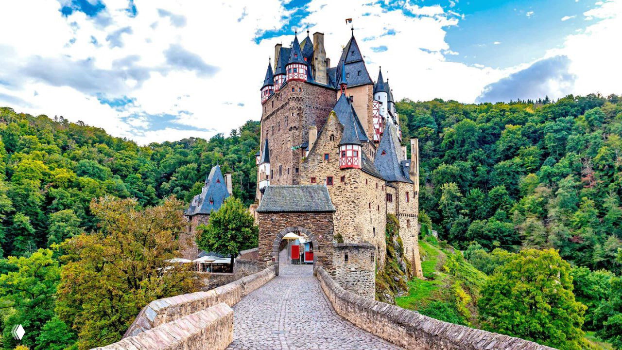 Подъездный каменный мост к замку Эльц, ведущий к воротам крепости на фоне зелёного леса и холмов — путь к исторической постройке.