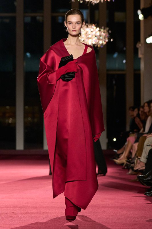 Модель в драпированном красном сатиновом плаще на подиуме Michael Kors в Метрополитен-опере; вечерняя атмосфера и акцент на силуэте.