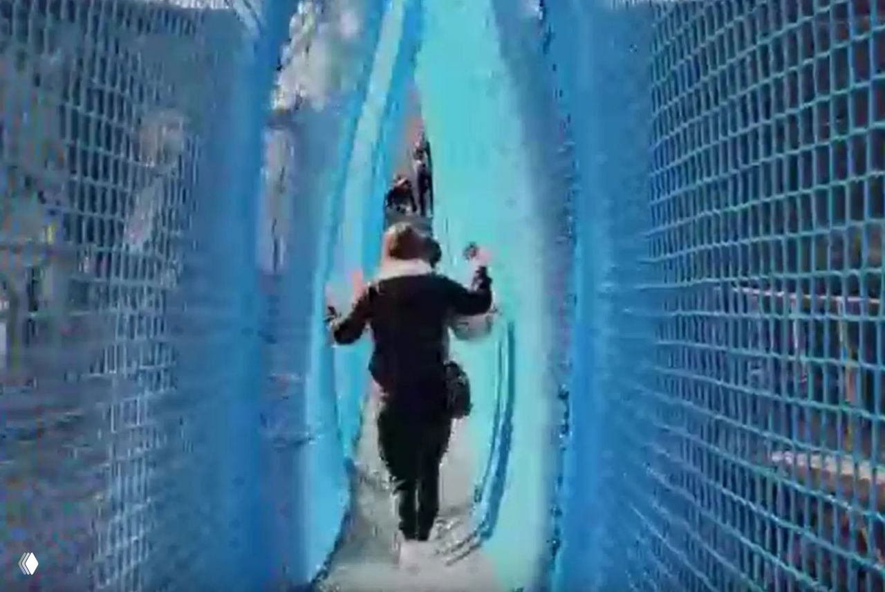 Проход внутри синей сетки подвесного моста: люди идут по туннелю на высоте между уровнями Эйфелевой башни, видна сетчатая конструкция.