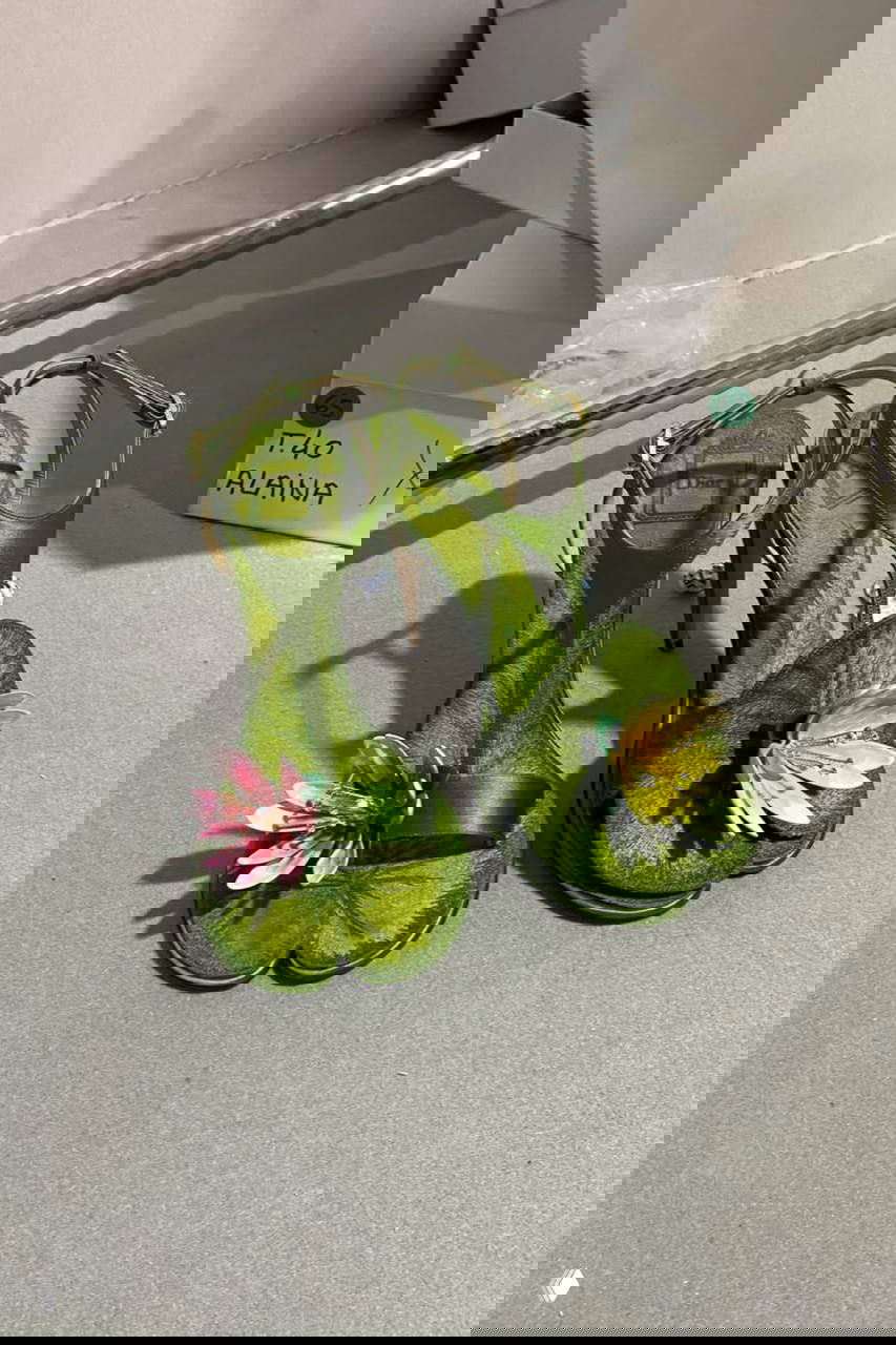 Зелёные босоножки на шпильке с объёмным цветком на носке, снятые на заднем фоне рабочего пространства — пример яркого аксессуара Dior.