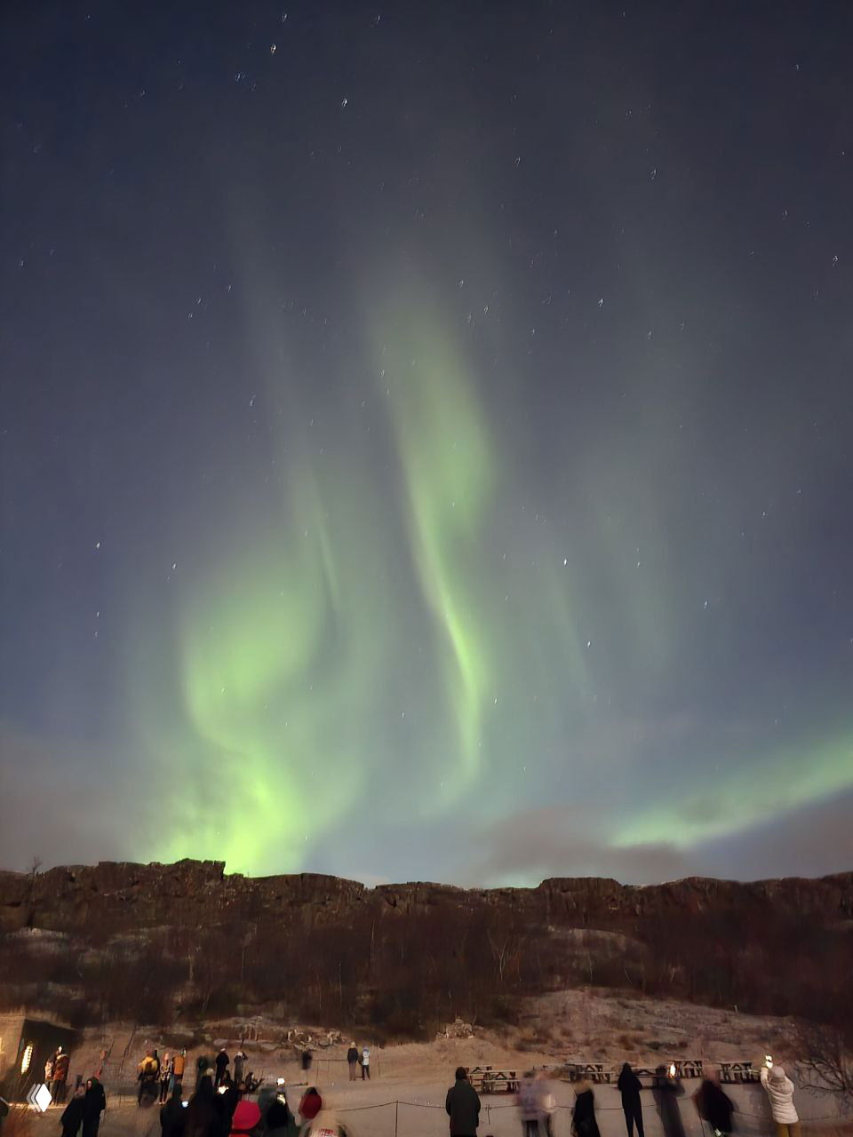 Вертикальные струи северного сияния над скалами; на переднем плане группа людей и палатки на заснеженной площадке, ночная сцена наблюдения природы.