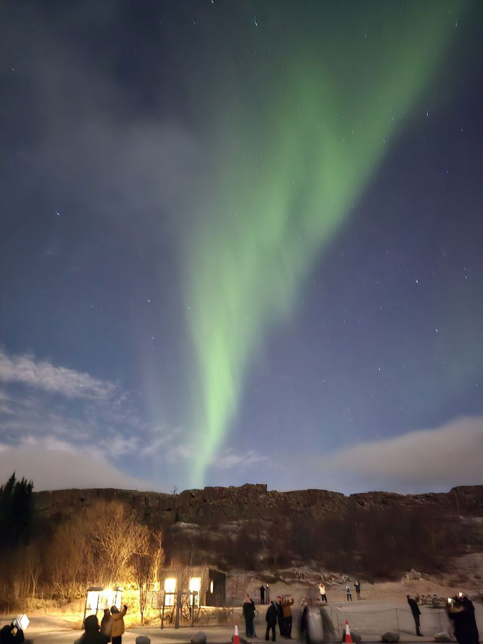 Высокая узкая зелёная полоса северного сияния, звёздное небо и группа наблюдателей у освещённых построек на переднем плане в тёмную ночь.