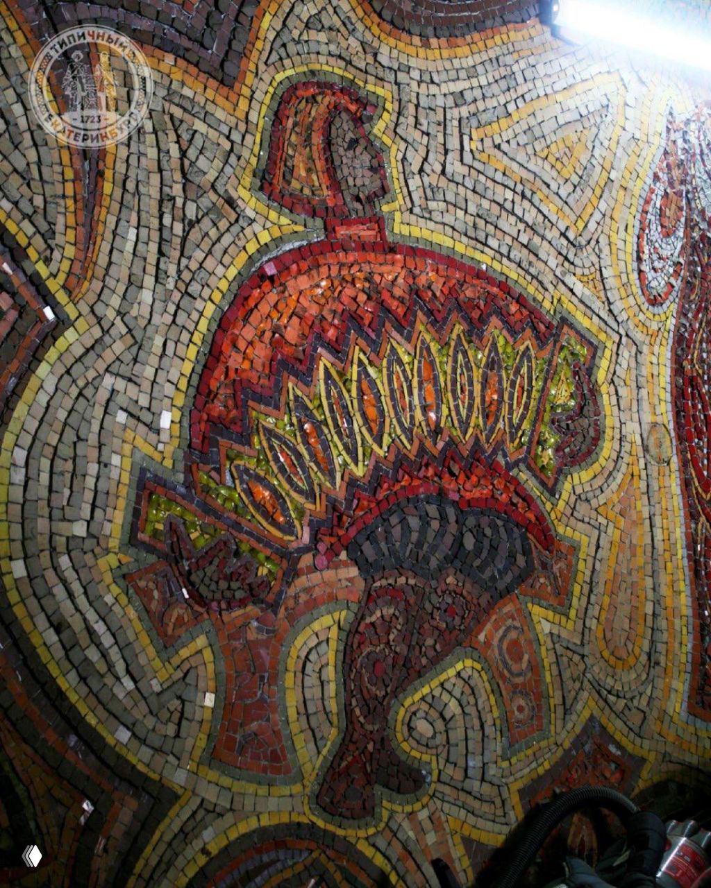 Фрагмент мозаики с орнаментом и фигурой, разноцветные смальты, видны швы и следы времени, часть мастерской на заднем плане.