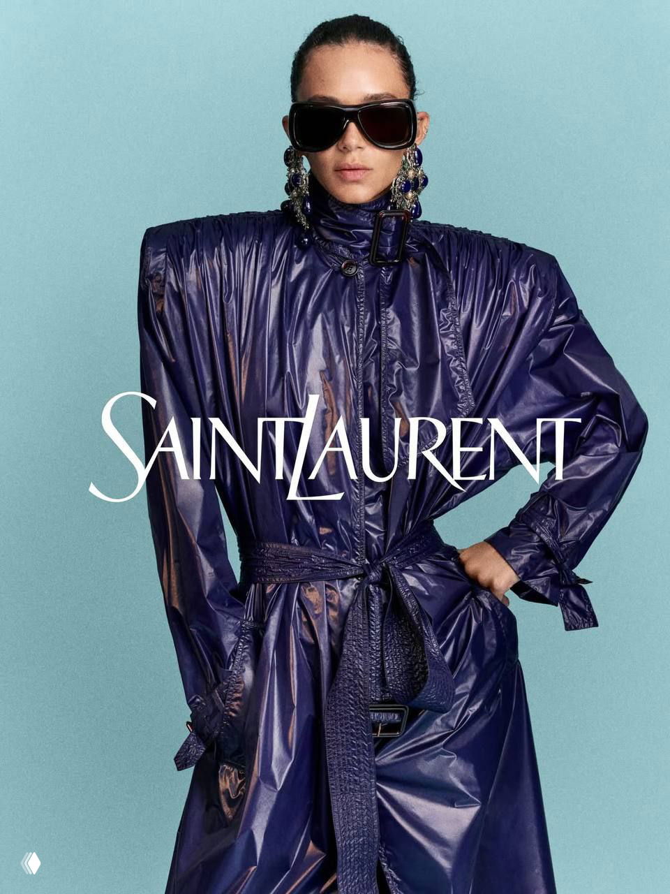 Фотография Saint Laurent в темно‑аметистовом оттенке: модель в блестящем плаще и больших солнечных очках на светлом фоне, строгий fashion‑образ.