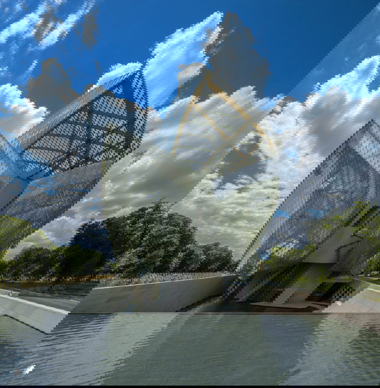 Крупный ракурс фасада резиденции Louis Vuitton: прозрачные стеклянные панели в виде парусов, архитектурные металлические и деревянные элементы на фоне неба.